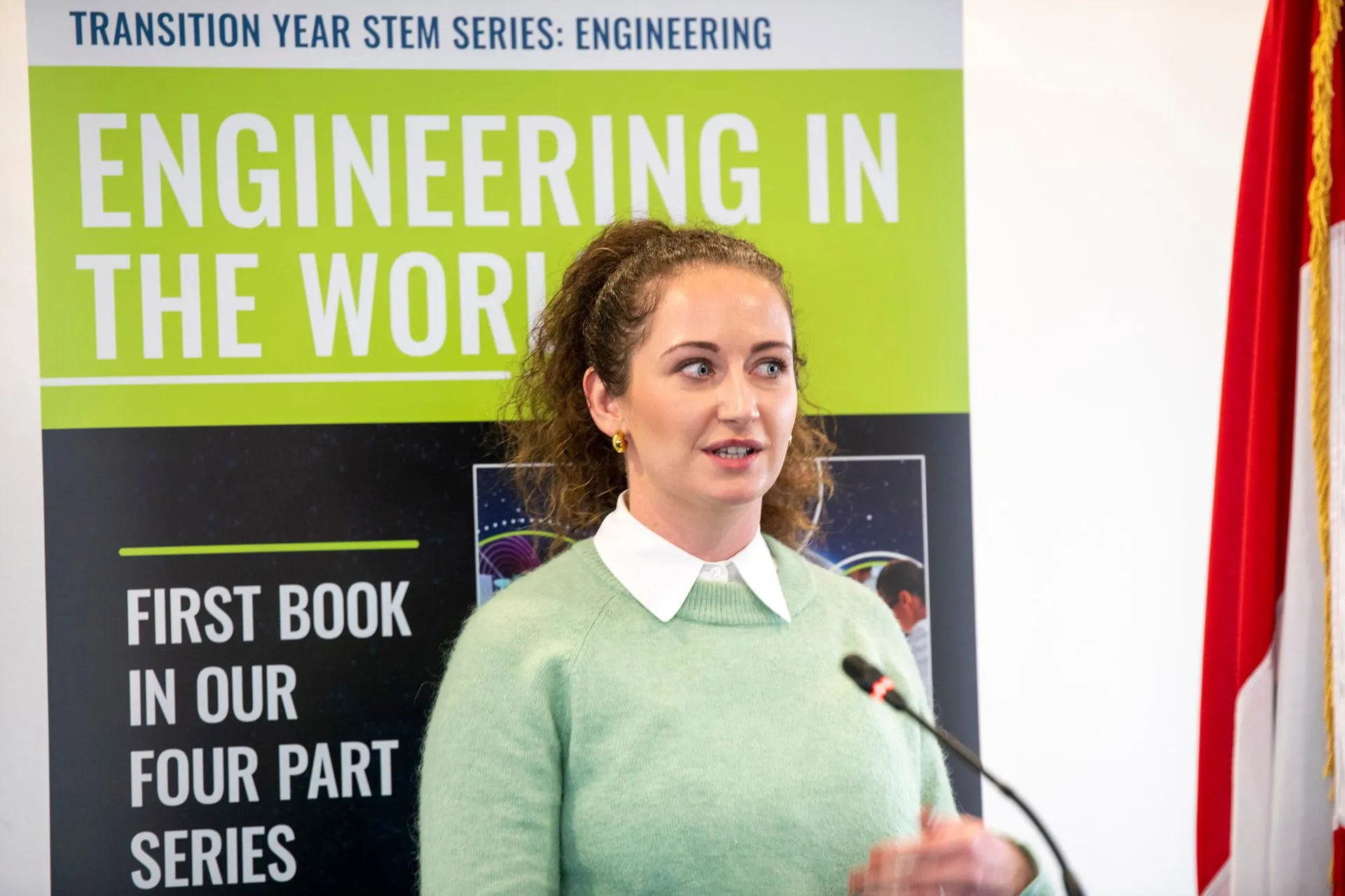 A woman in a green jumper speaking into a microphone in front of the book banner