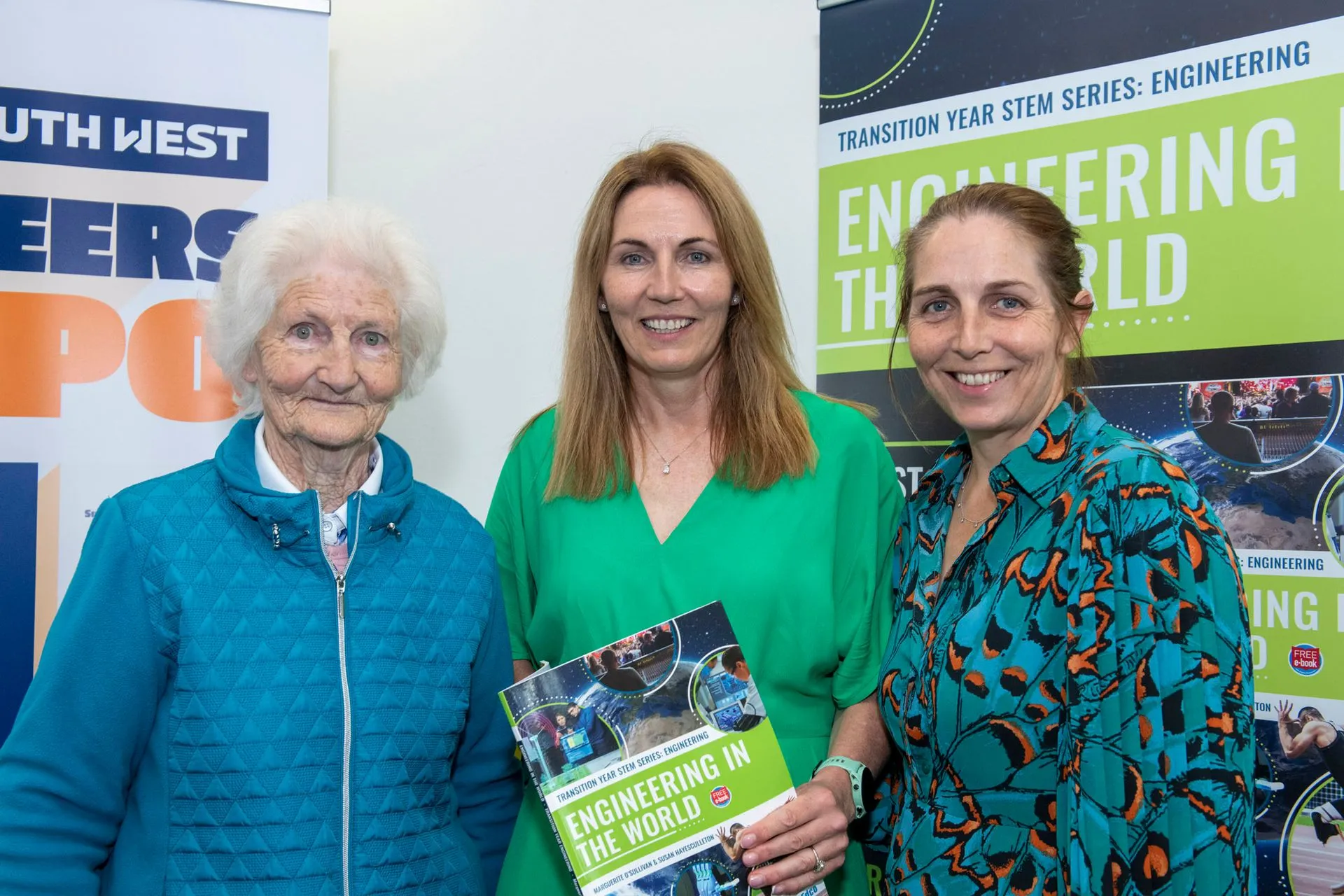 Three women holding copies of the book in front of the Engineering in the World and STEM South West Careers Expo banners